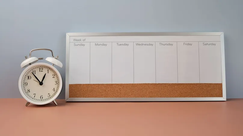 A white alarm clock next to a weekly organiser whiteboard.