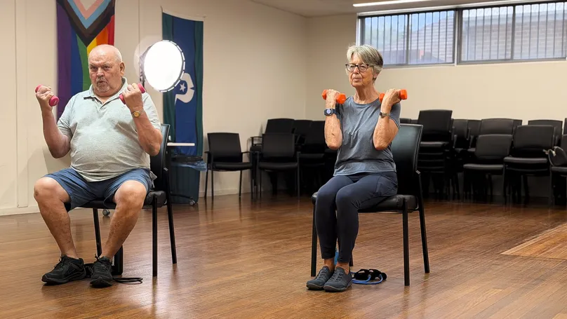 Two older people demonstrating seated biceps curls with dumbbells.