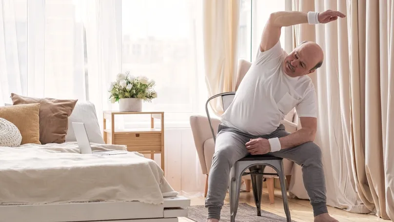 An older man doing chair yoga poses in his bedroom, following along with a video playing through the laptop on his bed.