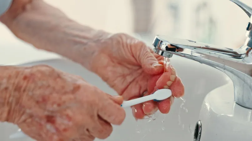 An older person's hands using a toothbrush to clean dentures under the running tap.
