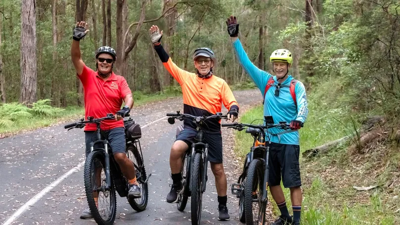 Older group of individuals in bright - coloured t-shirts and helmets, smile as they wave to the camera, whilst posing with their bicycles.
