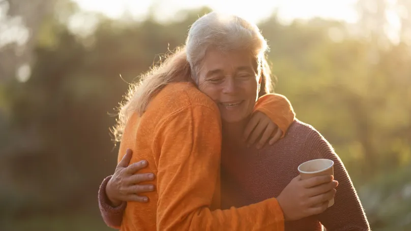 An older woman embracing a younger woman with greenery in the background.