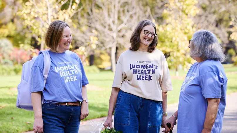 3 women in purple Women's Health Week shirts smile and chat with each other at the park.