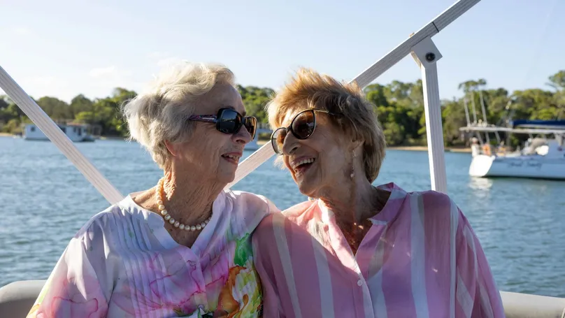 Two smiling women stand arm in arm beside the water at sunset, wearing colourful pink clothing and looking toward the camera.