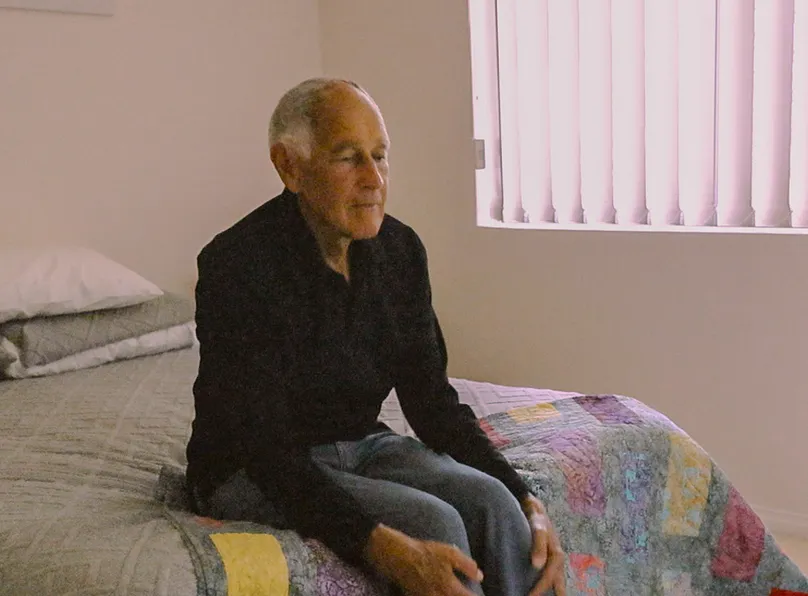 Older man in bedroom sitting on bed.