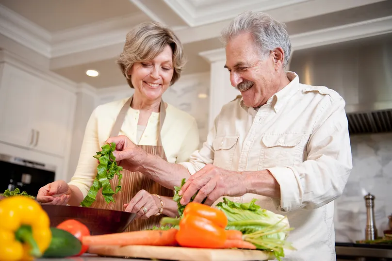An older couple smiling and preparing vegetables in the kitchen.