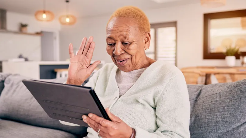 An older woman smiling and waving at someone through video call on her iPad.