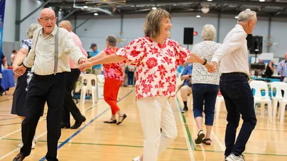 A group of older people holding hands and dancing in an indoor sports hall