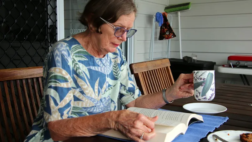 Woman with dark blonde hair wearing glasses and a patterned dress, reads a book as she enjoys a cup of tea outdoors.