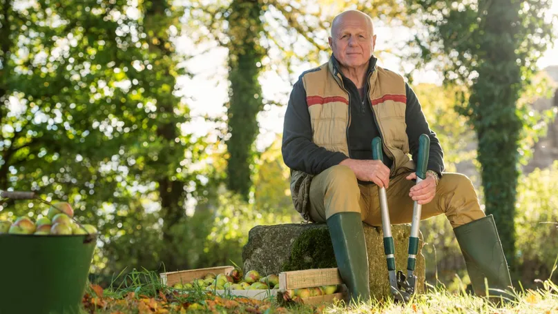 A man sitting on a seat in the garden with mid-length pruners. The pruners allow him to sit while reaching the ground without stooping.