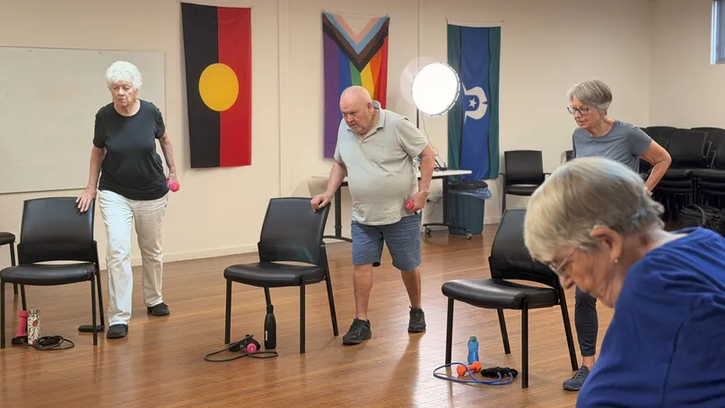 Older people standing while holding dumbbells in one hand, in the first position of the triceps kickback.