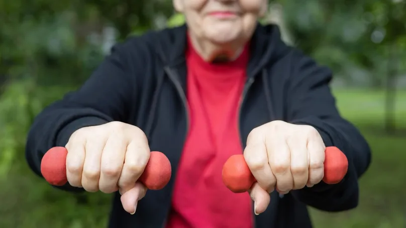 An older woman holding up a dumbbell in each hand while exercising in the park.