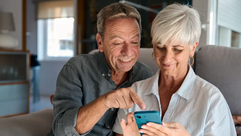 An older couple smiling, both looking at the same smartphone.