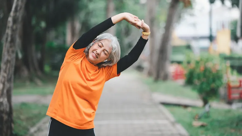 An older woman wearing a bright orange shirt stretching her arms overhead in a park.