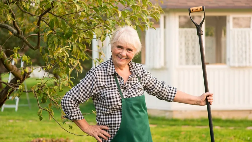 An older woman gardening with a long-handled shovel. The tool is taller than she is, so she can dig while fully upright.