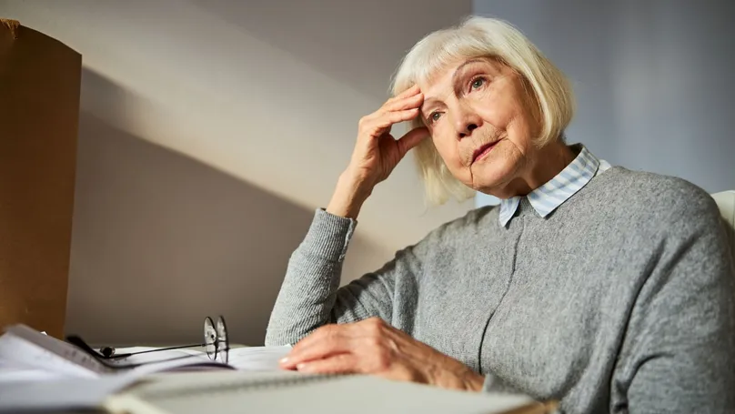 An older woman rubs her temple and stares into the distance while sitting at her desk.