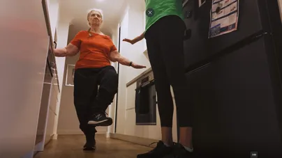 Kamilla Haufort and an older woman demonstrating tandem stance in the kitchen.