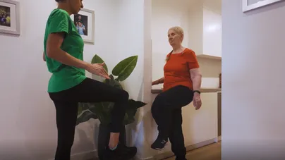 Kamilla Haufort demonstrating standing leg exercises alongside an older woman in the kitchen.